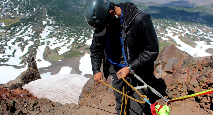 Wearing safety gear and secured by ropes, a person stands on the edge of a cliff, high above a landscape dotted with snow.